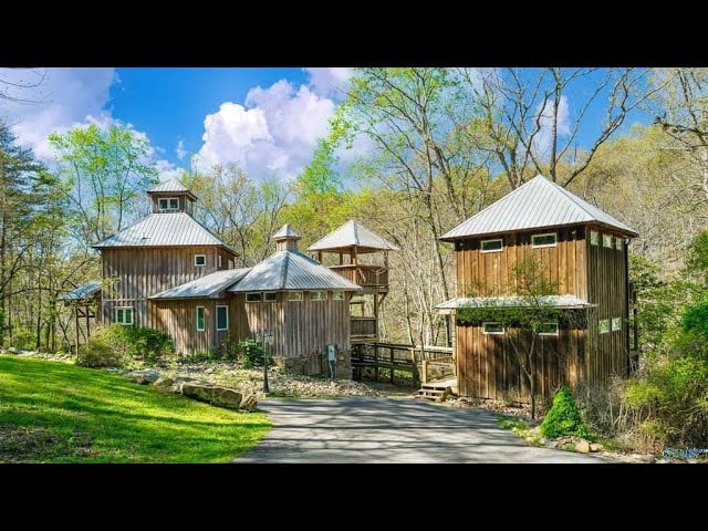 Rustic wooden buildings with metal roofs beside a paved drive and green lawn.