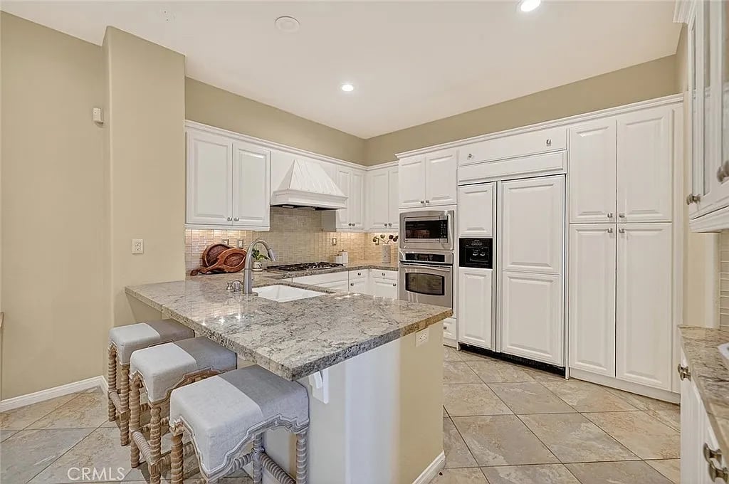 Kitchen with a large stone island, three upholstered stools, white cabinetry, and stainless steel appliances.