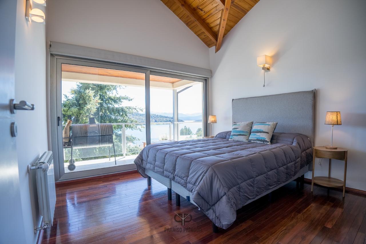 Bedroom with a gray quilted bed, vaulted timber ceiling, and balcony overlooking a lake.