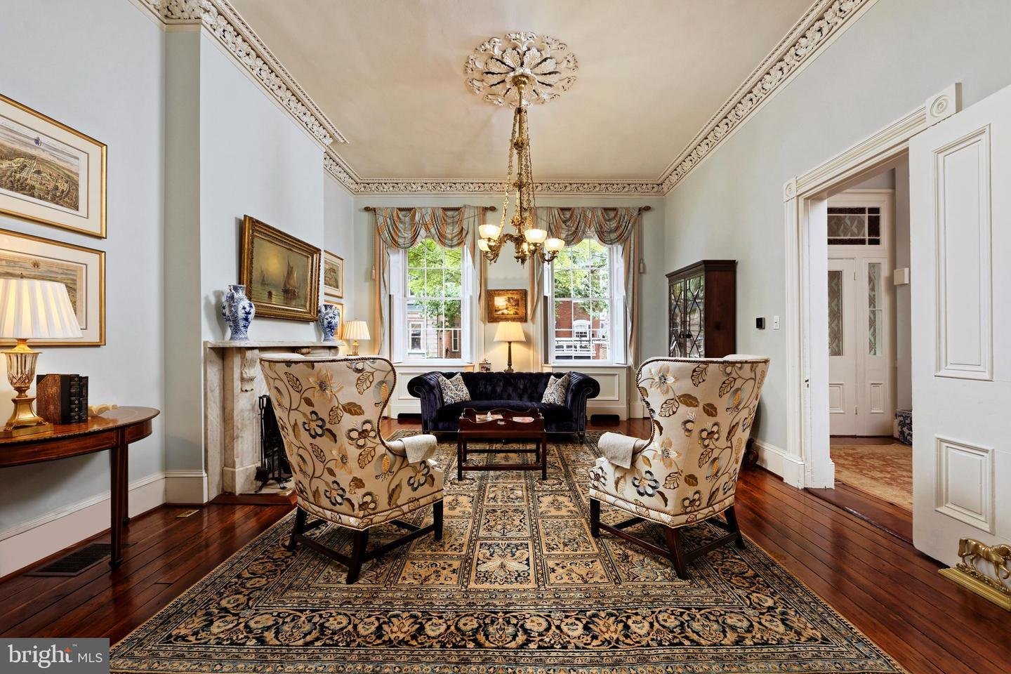 Living room with ornate ceiling trim, chandelier, fireplace, and tall windows.