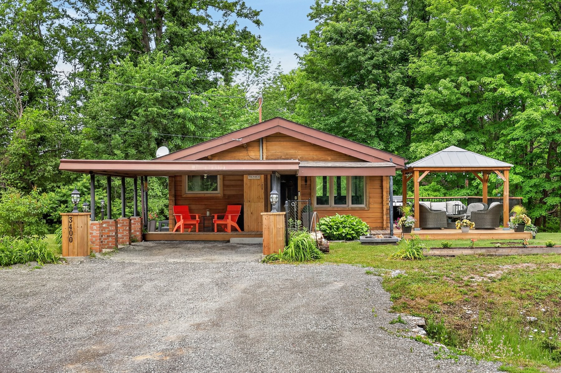 Wood-sided cottage with a covered porch, red chairs, gravel driveway, and furnished gazebo among leafy trees.