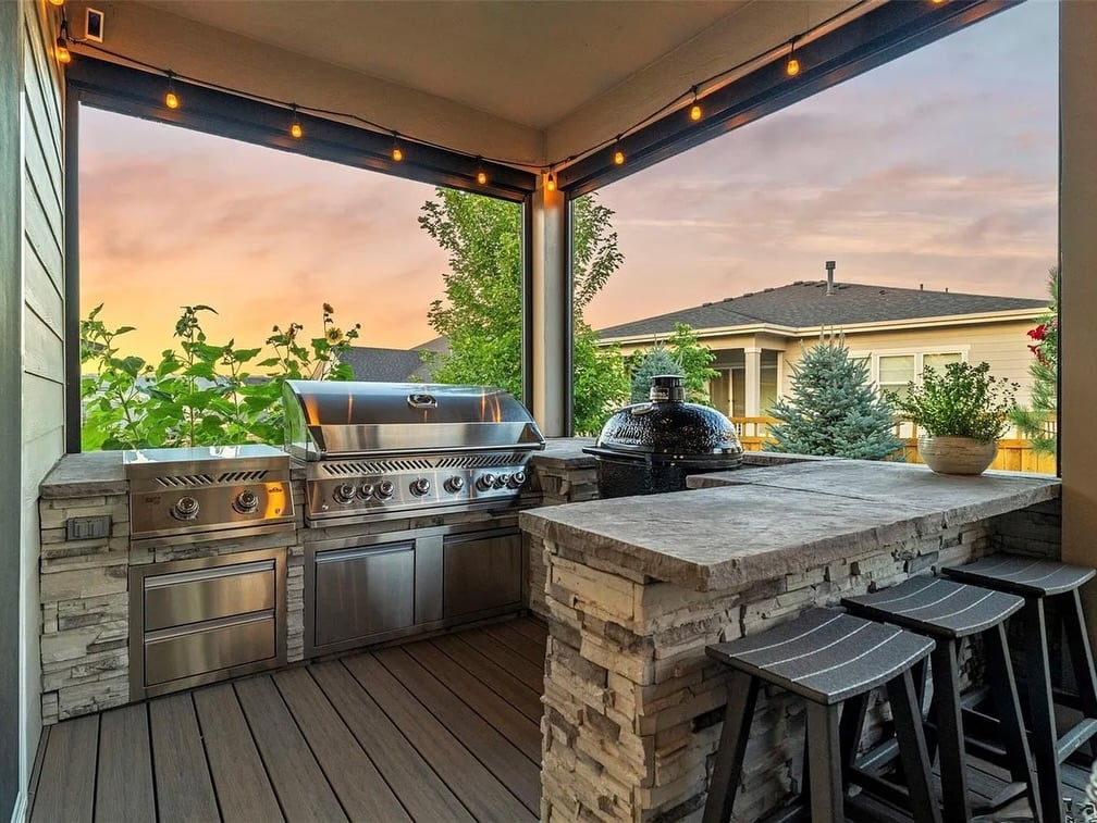 Ranch-style home backyard patio in The Meadows, Castle Rock, with custom outdoor kitchen featuring grill, sizzle zone, and smoker, surrounded by mature trees and twinkling lights—designed for gatherings and entertaining.