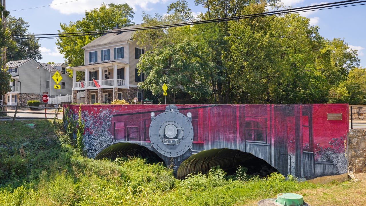 Train mural numbered 571 on a small bridge beside a leafy residential street.