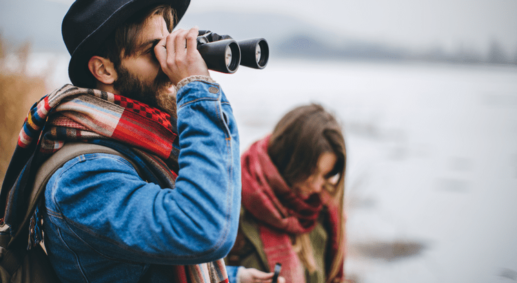 A man in a hat uses binoculars near a serene, misty lake, with a woman focused on her smartphone beside him. Both are wrapped in colorful scarves.