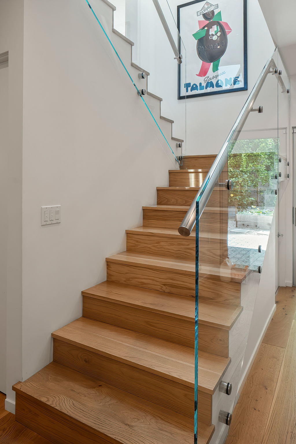 Oak staircase with glass railing and framed artwork, overlooking a garden.