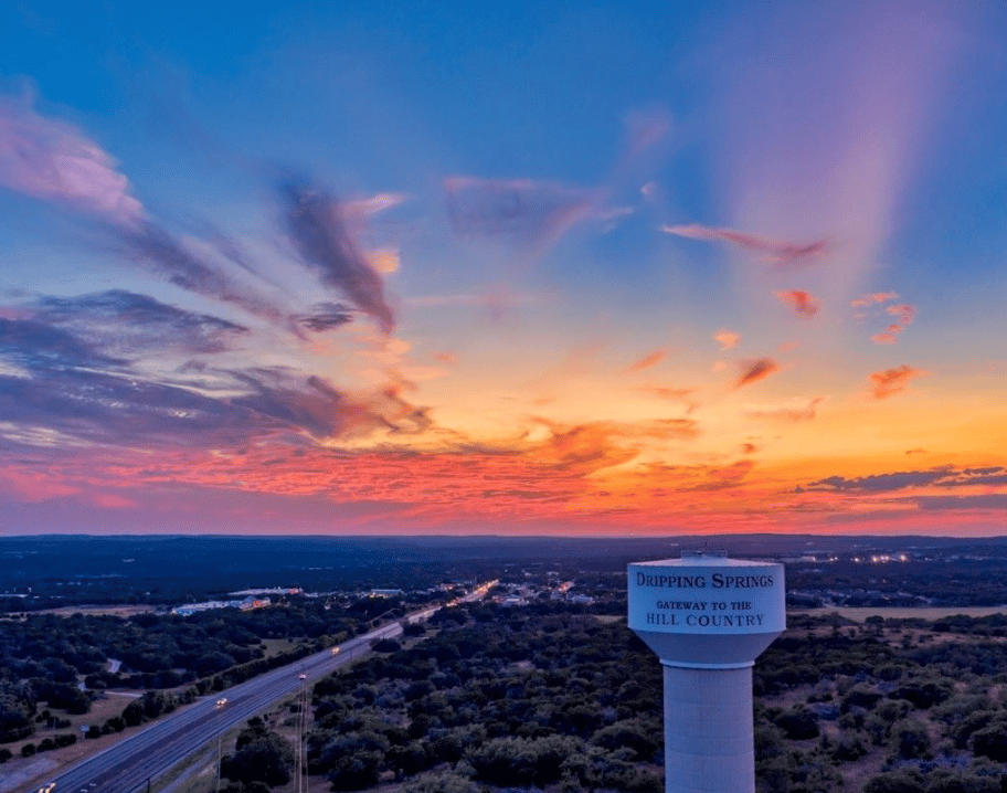 Dripping Springs water tower above the town and highway beneath a colorful sunset.