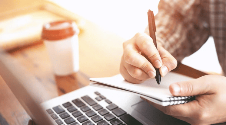 Person writing in a notebook beside a laptop and takeaway coffee.