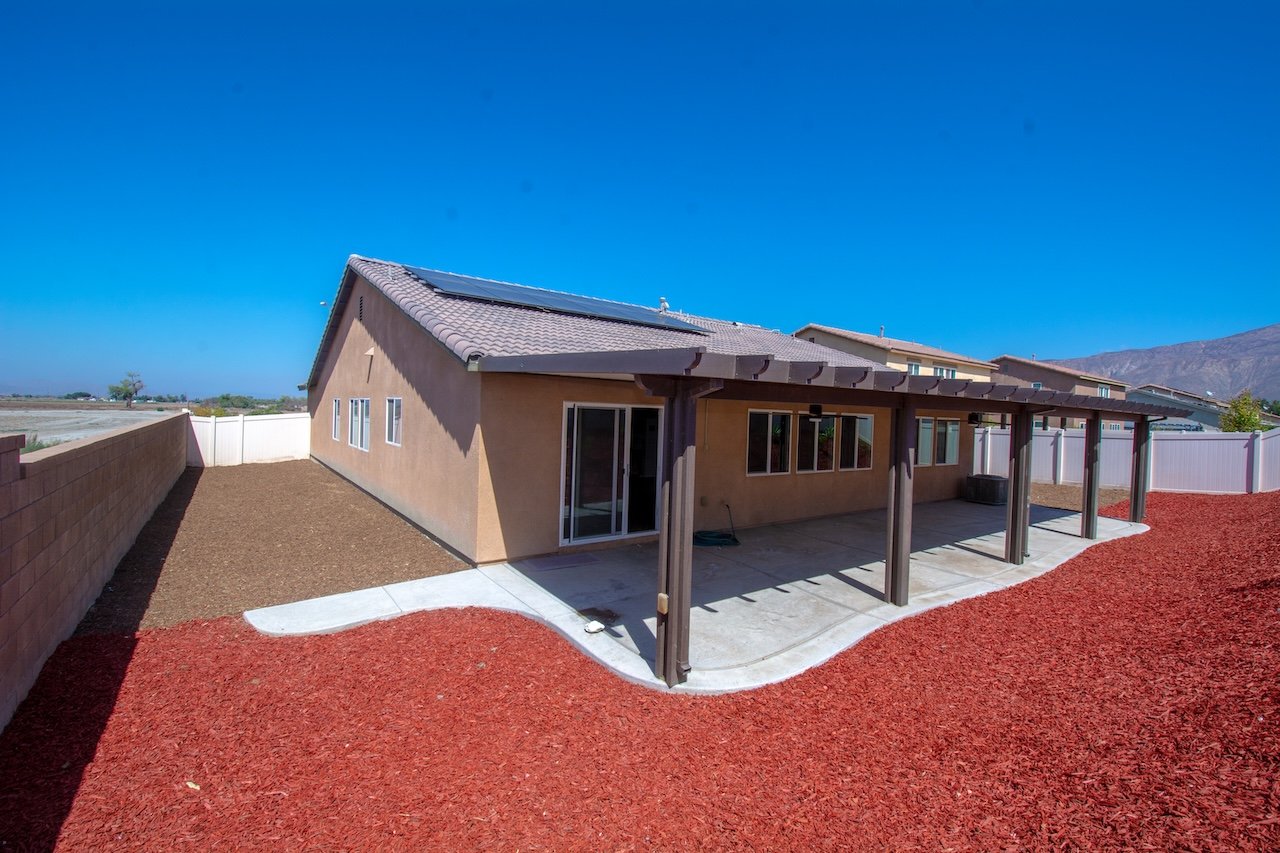 Rear of a stucco house with rooftop solar panels, a covered patio, and red mulch landscaping.