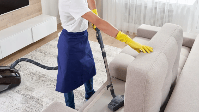 Cleaner vacuuming beside a light sofa in a bright living room.