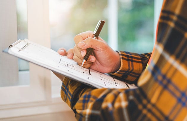 Person in a plaid shirt writes on a home inspection checklist beside a window.
