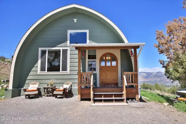 Green arched-roof home with a wooden porch, outdoor seating, and mountain views.