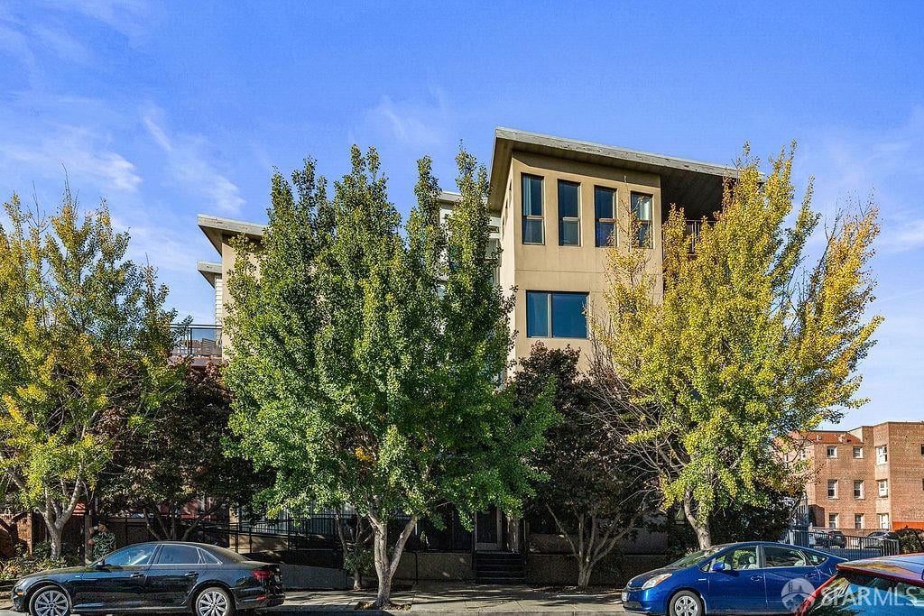 Modern multi-story residential building partly obscured by leafy trees, with parked cars along the street.