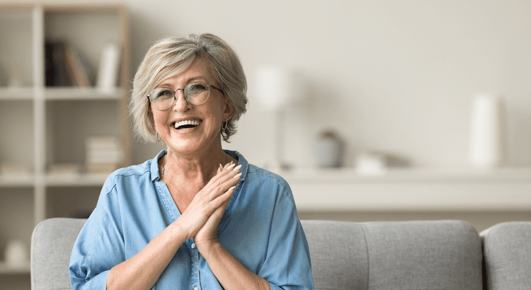 An elderly woman smiling warmly, wearing glasses and a light blue top.