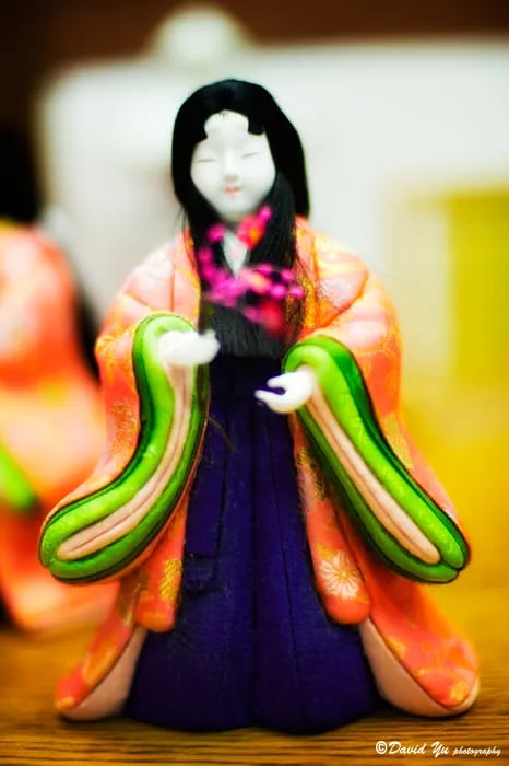 A close-up of a traditional Japanese doll wearing a colorful orange, green, and purple kimono, holding a small branch of pink flowers.