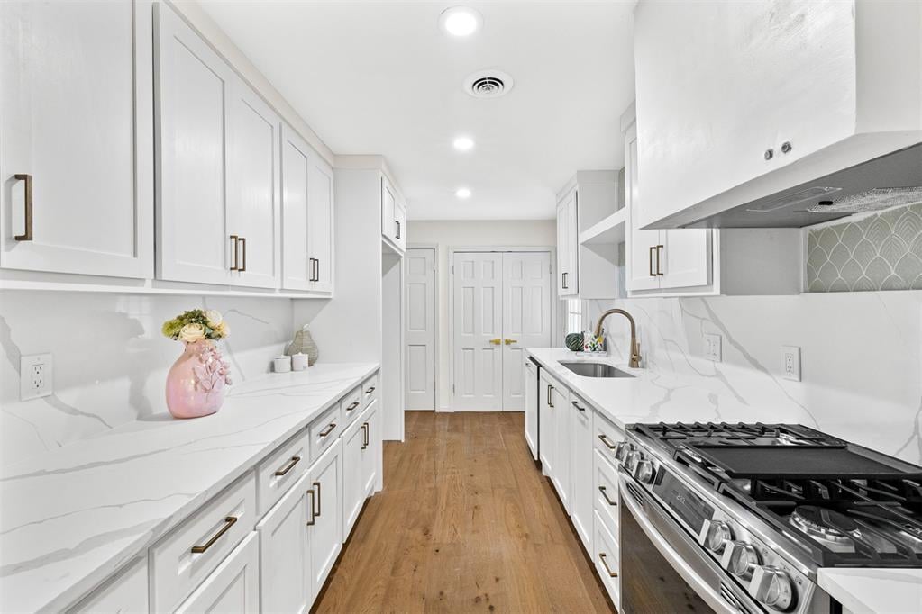 Bright white galley kitchen with marble-look counters, brass hardware, stainless steel range, and warm wood flooring.