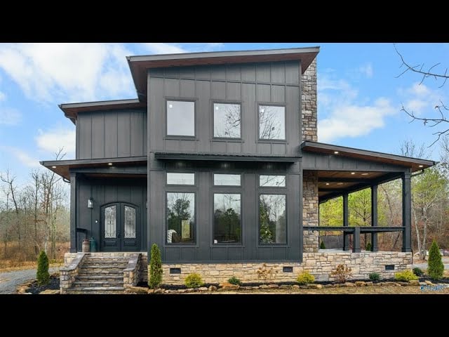Modern dark-sided two-story home with a stone chimney, broad windows, and a covered porch.