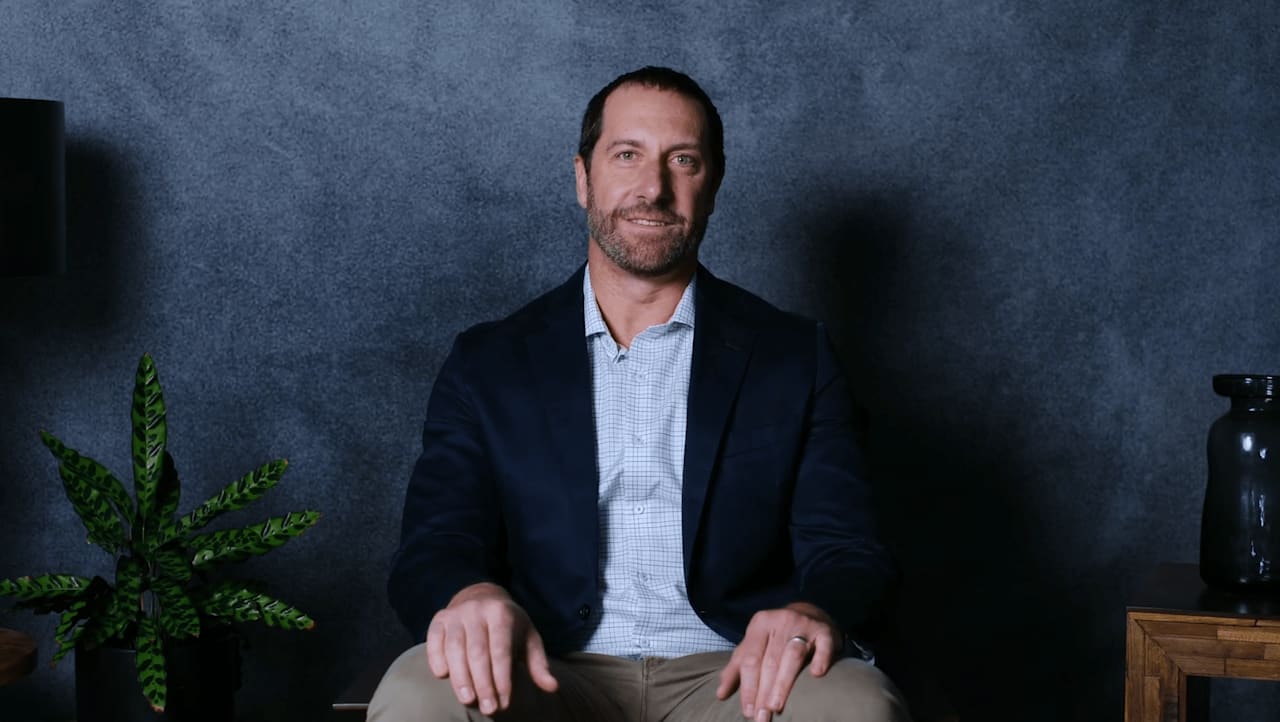 Seated man in a navy blazer against a dark textured wall, with a leafy plant beside him.