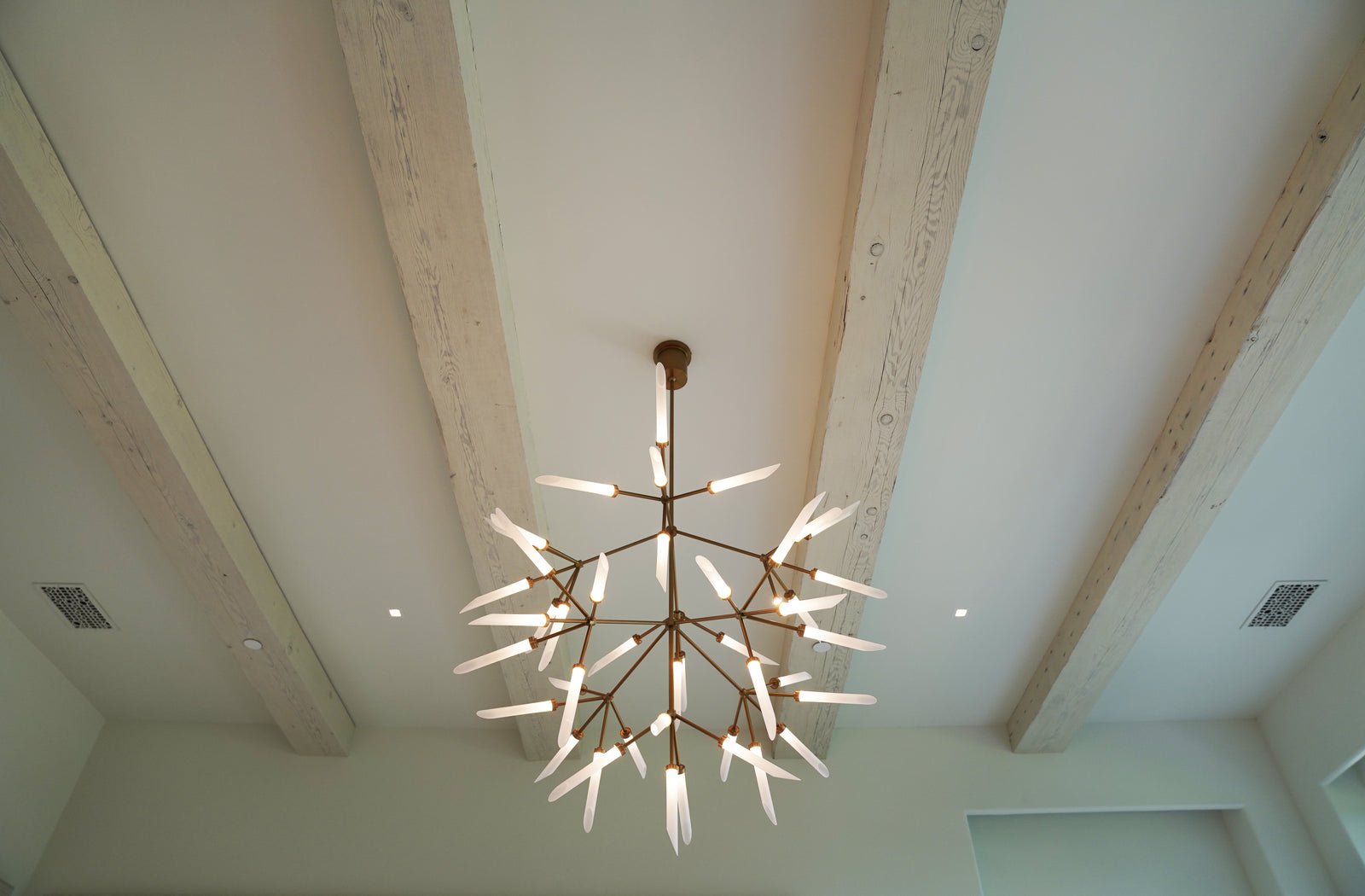 Looking up at a white ceiling crossed by whitewashed wood beams, with a branching glass chandelier and smooth ceiling panels between the beams.