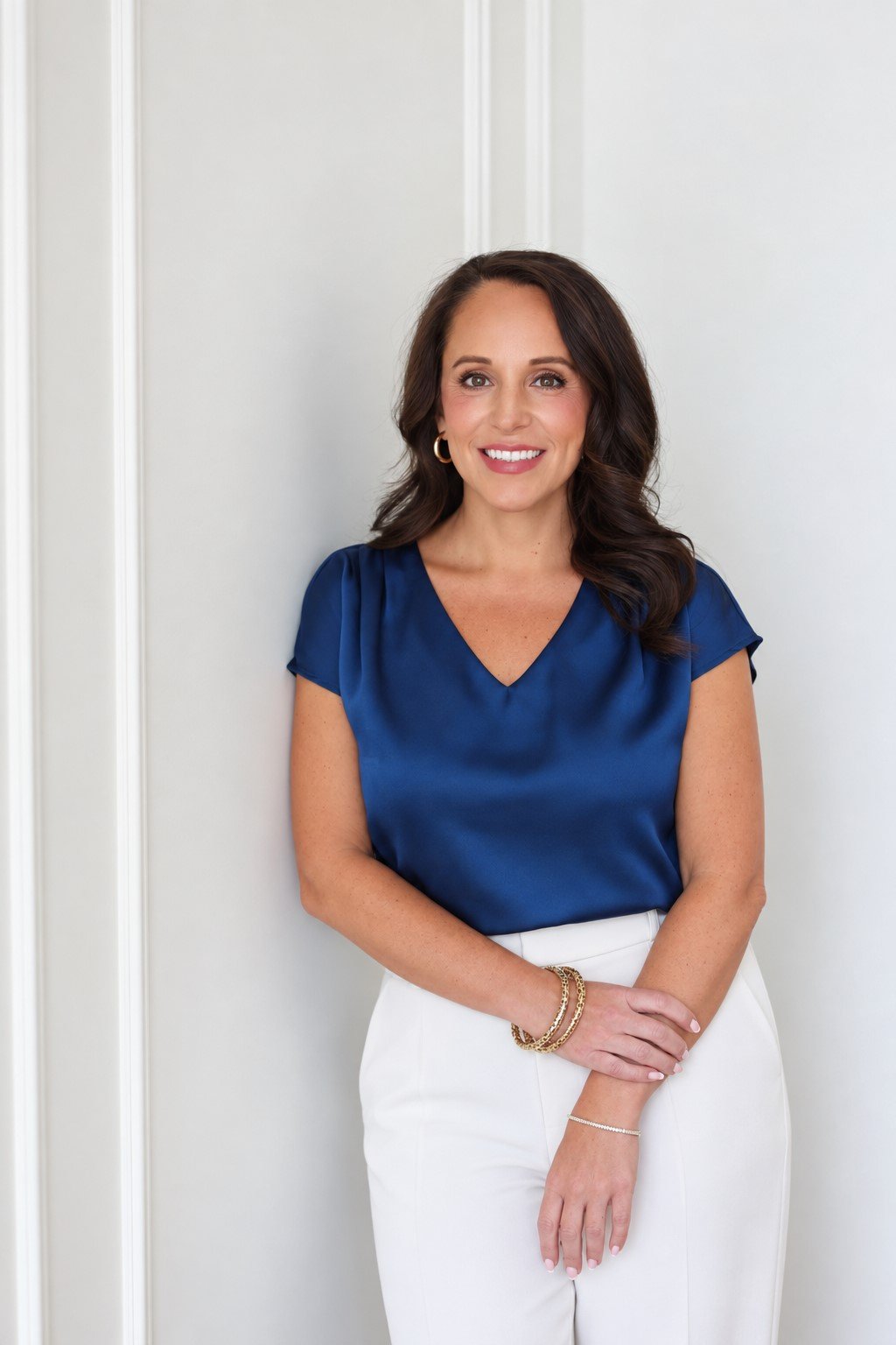 Traci James smiling in a navy blouse and white pants, posed against a light paneled wall.