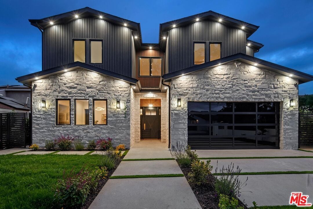 Modern two-story stone-and-siding home at dusk with illuminated facade and black garage door.