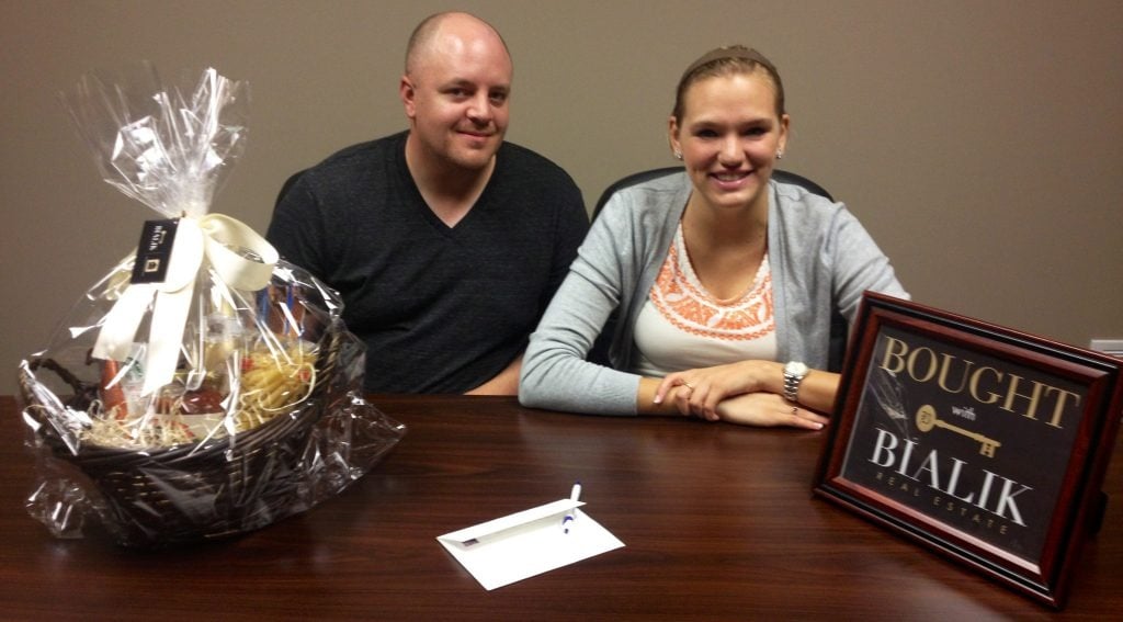 Smiling couple celebrates their home purchase beside a Bialik Real Estate sign and gift basket.