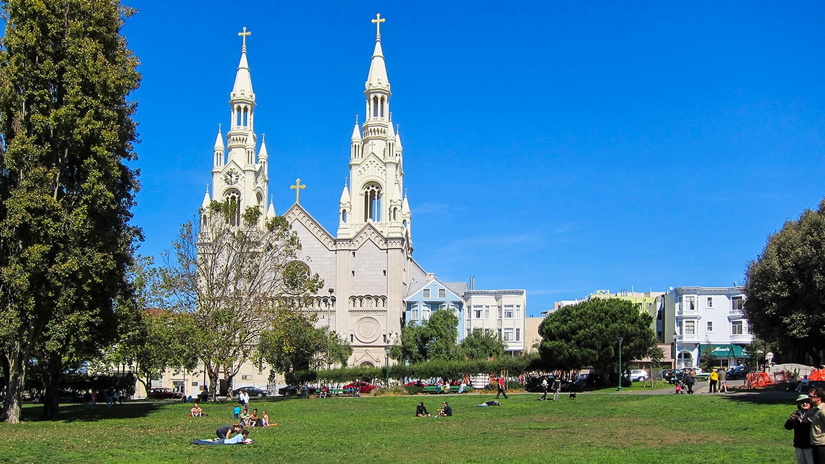 Daytime view of Washington Square Park in San Francisco, featuring a lush green lawn with people relaxing and enjoying the sunny weather. The iconic Saints Peter and Paul Church stands prominently in the background against a clear blue sky, surrounded by trees and residential buildings.