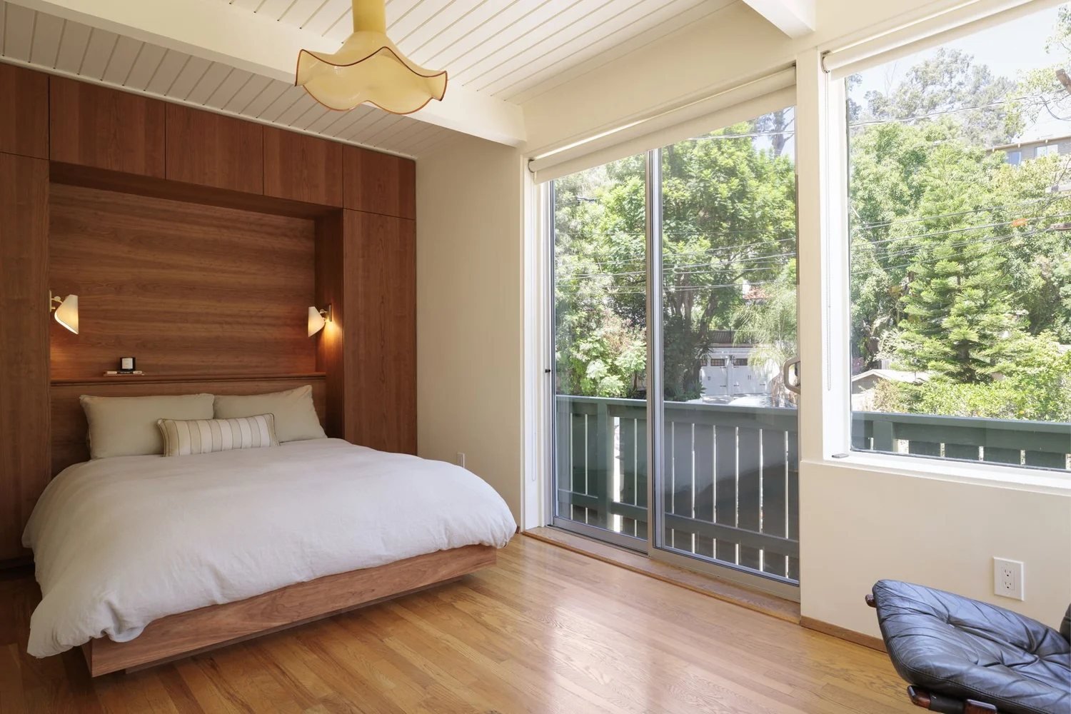 The primary bedroom with a warm wood headboard wall, a low platform bed, and sliding glass doors opening to a balcony above the trees.