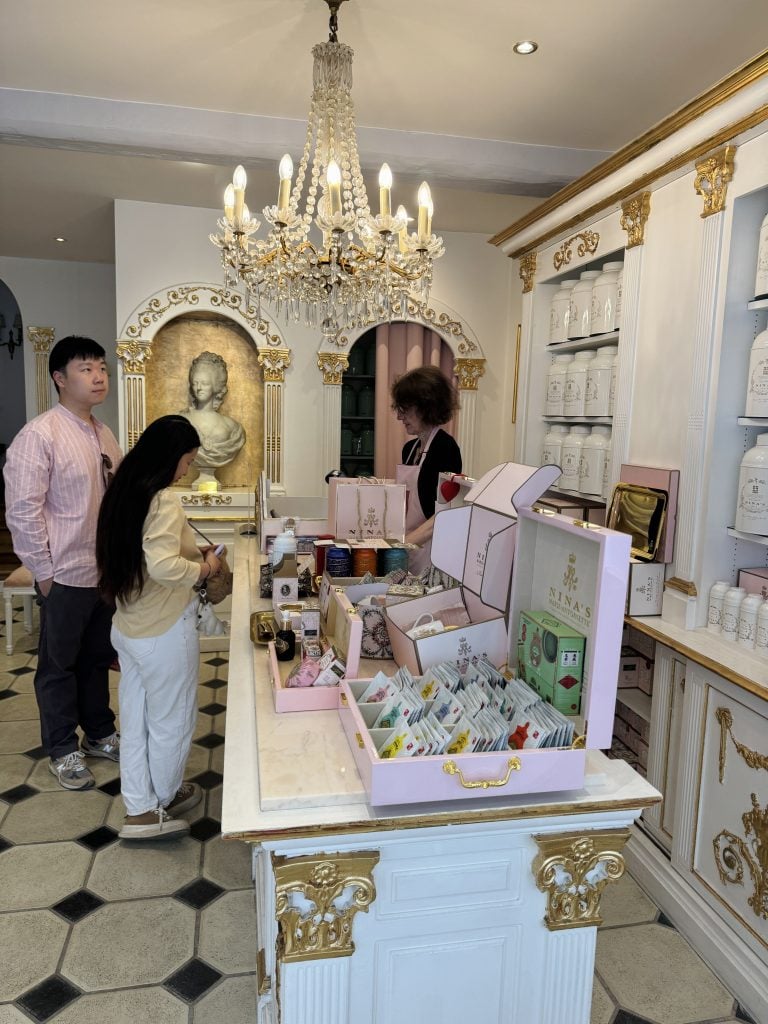 Ornate boutique interior with a crystal chandelier, gilded cabinetry, and pink cosmetics displays.