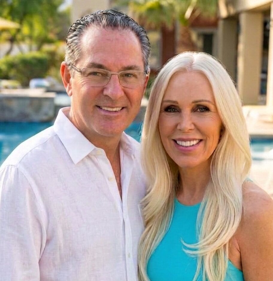 Two smiling real estate agents pose beside a pool; he wears glasses, and she wears a turquoise top.