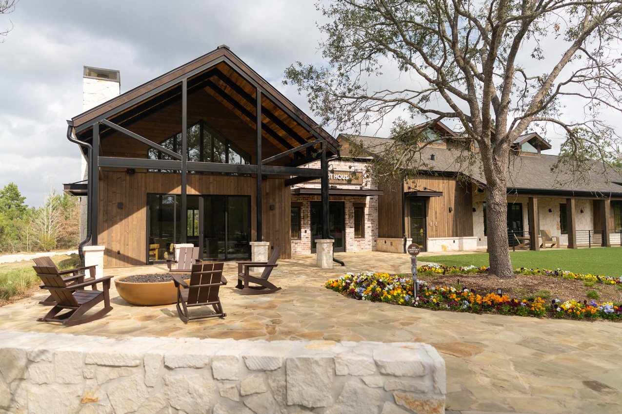 Community clubhouse with a timber-and-stone facade, stone patio, outdoor chairs, and flower beds.