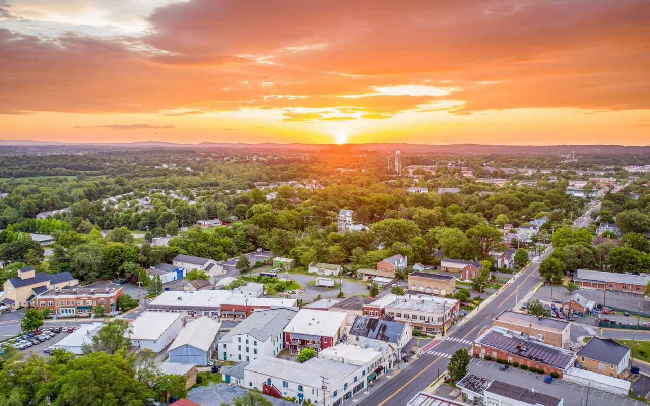 Aerial view of a small town at sunset, with leafy neighborhoods, low-rise shops, and wooded hills.
