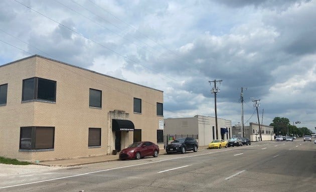 Tan brick commercial building with dark windows beside a street lined with parked cars.