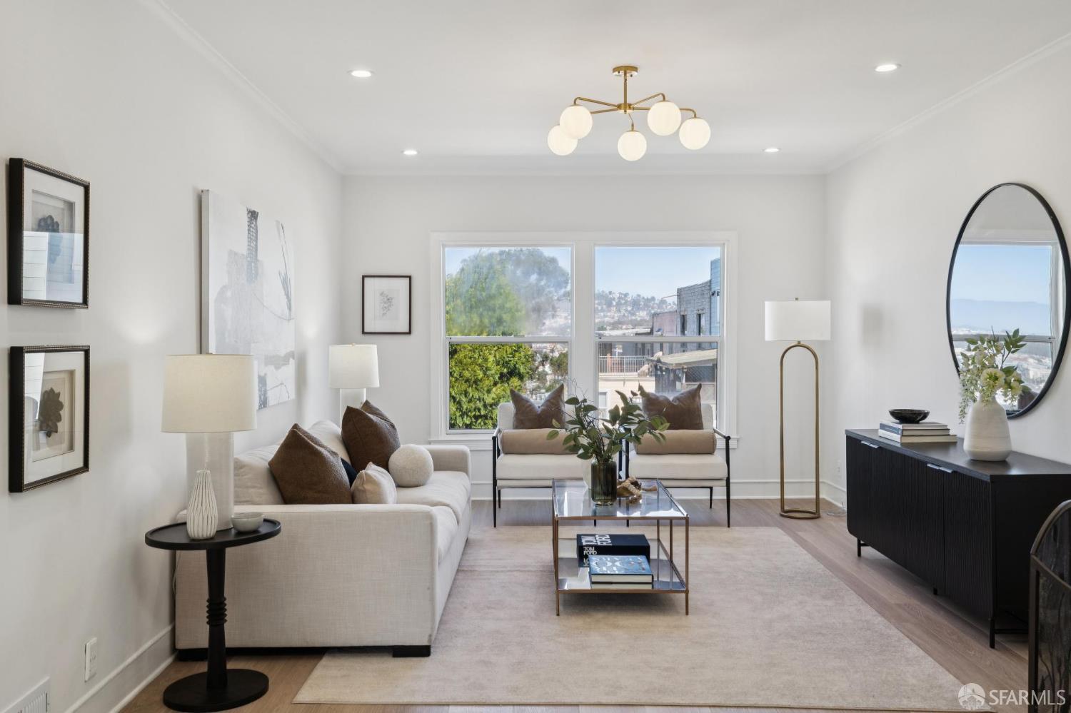 Staged living room with a cream sectional, glass coffee table, and large windows overlooking nearby buildings and hills.