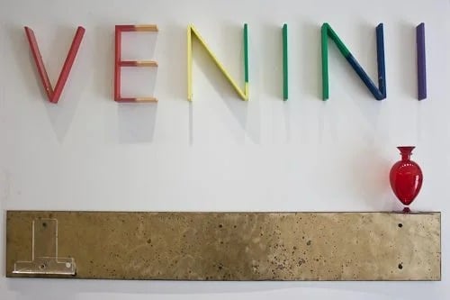 Colorful VENINI glass letters above a tan stone panel, with a small red glass vase.