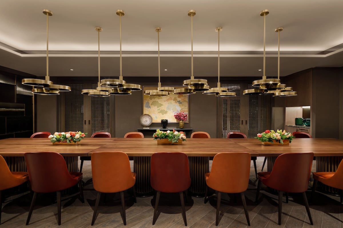 Communal dining room with a long wooden table, orange chairs, and brass pendant lights.