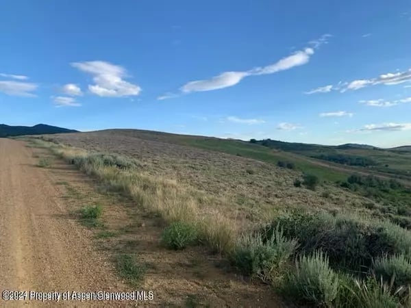 Rolling sagebrush-covered hills beside a dirt track under a blue, cloud-dotted sky.