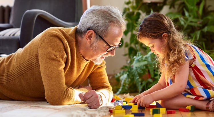 An elderly man, dressed in a mustard-colored sweater, engaging with a young girl. They are both involved in building with colorful blocks.