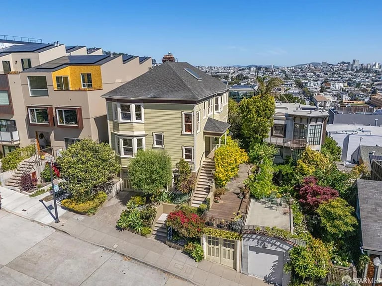 Pale green Victorian-style house with a landscaped front garden, hillside homes, and San Francisco in the distance.