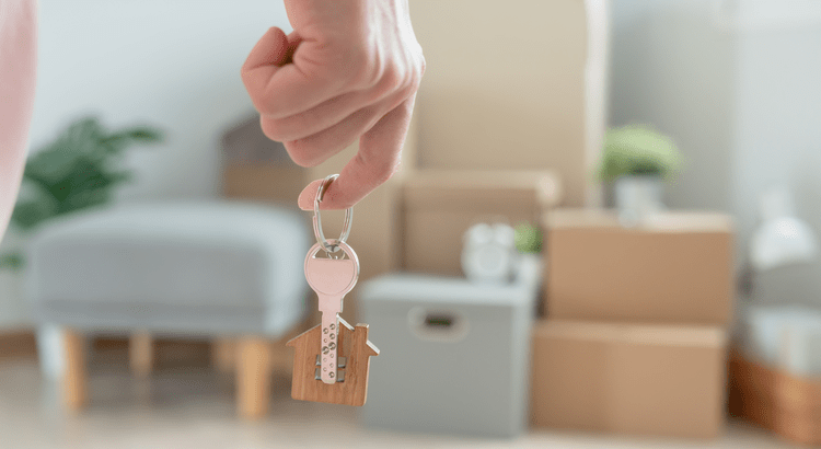 A hand holds a house-shaped keychain with boxes and a chair blurred in the background, suggesting moving or new home ownership. The tone is hopeful and welcoming.