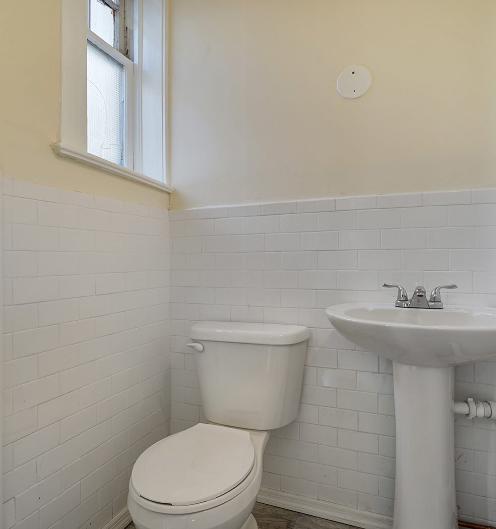 Bathroom with white tiled walls, a toilet, pedestal sink, and high window.