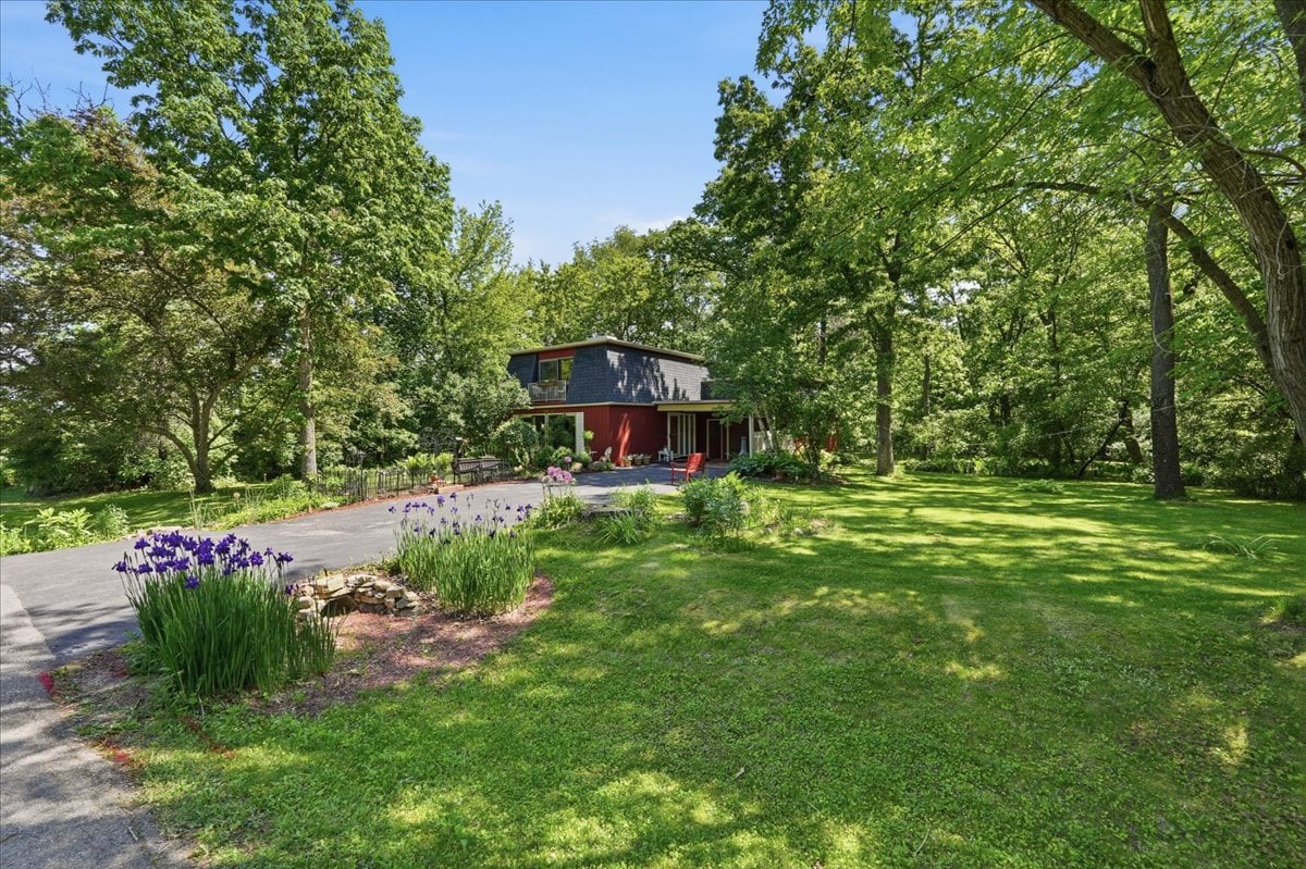 The exterior of 7062 Craig Lane in the Sherwood Forest neighborhood near Sun Prairie, Wisconsin, showing the home surrounded by mature trees, perennial gardens, and a sweeping wooded lot on just shy of an acre.