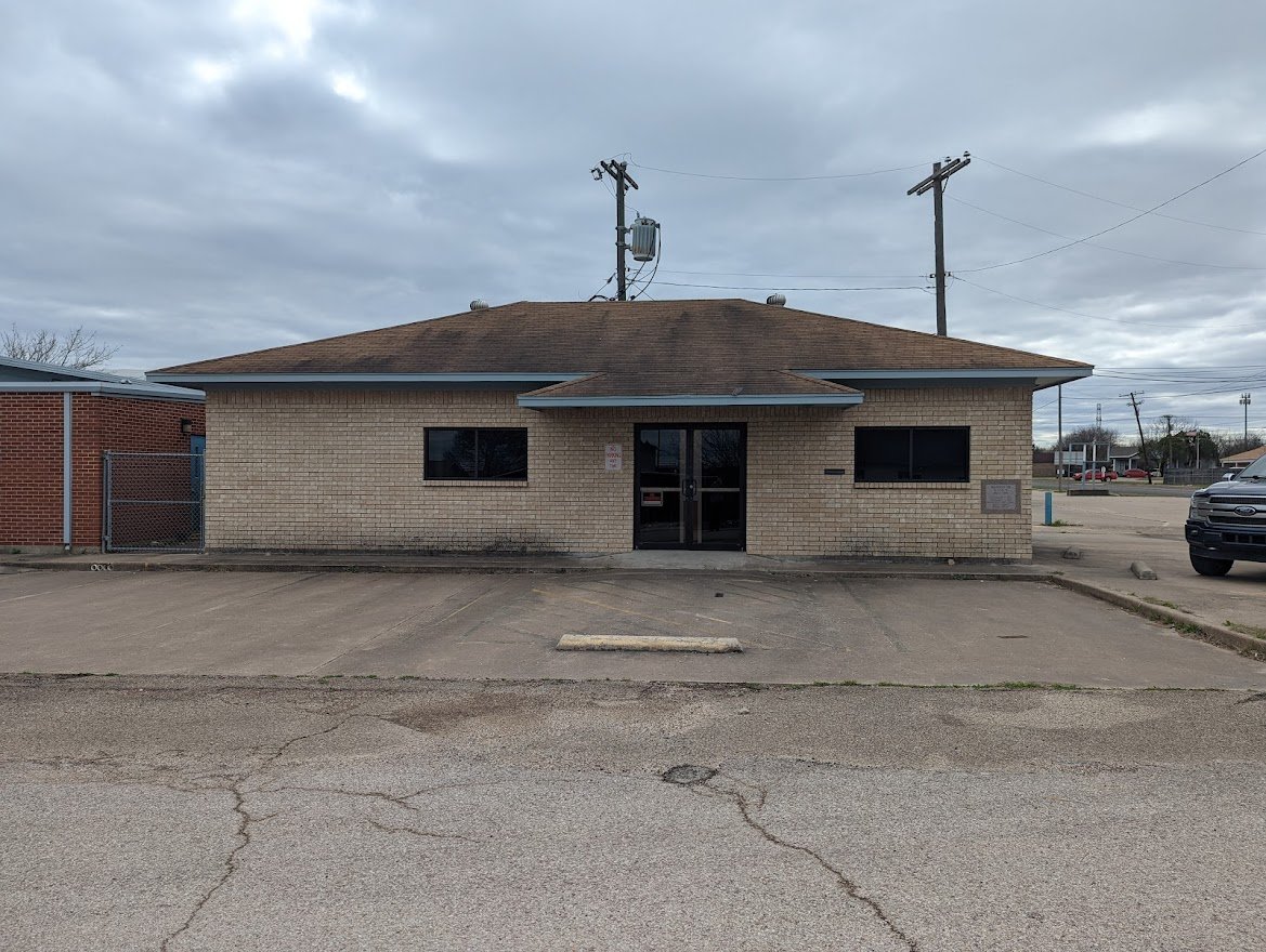 Single-story tan brick commercial building with a centered glass entrance and paved parking area.