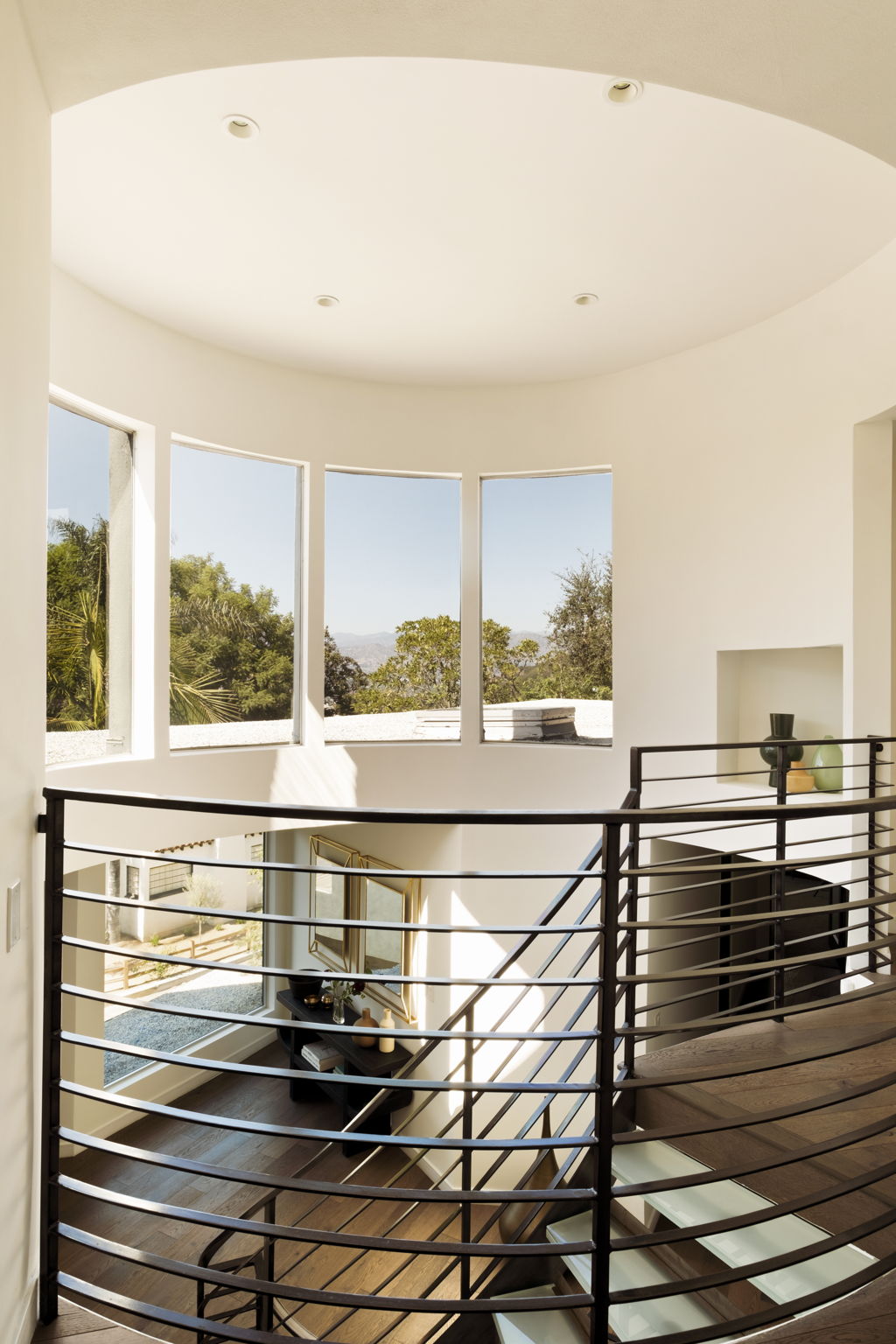 Curved stairwell with dark metal railings, wood steps, and tall windows overlooking trees and hills.