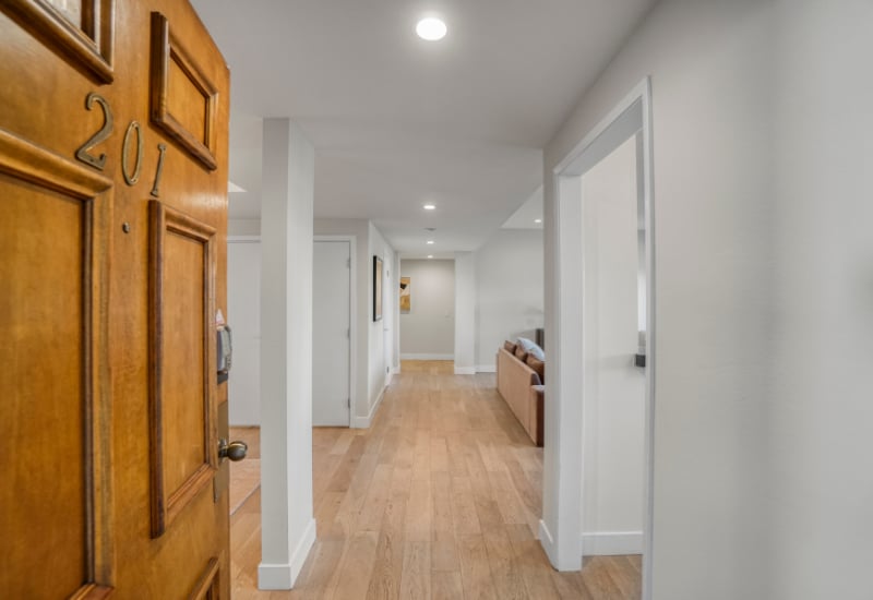 Apartment hallway with light hardwood floors, viewed past a paneled wooden entry door.