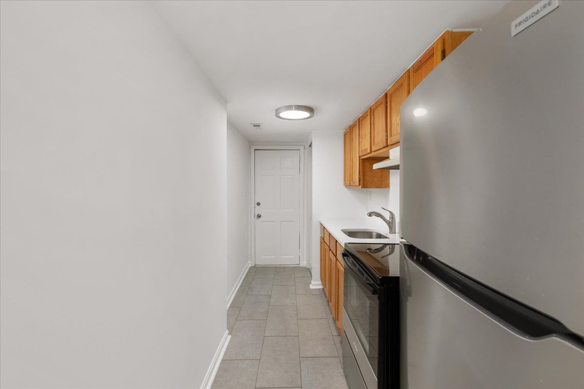 Narrow galley kitchen with wood cabinets, stainless steel refrigerator, and gray tile flooring.