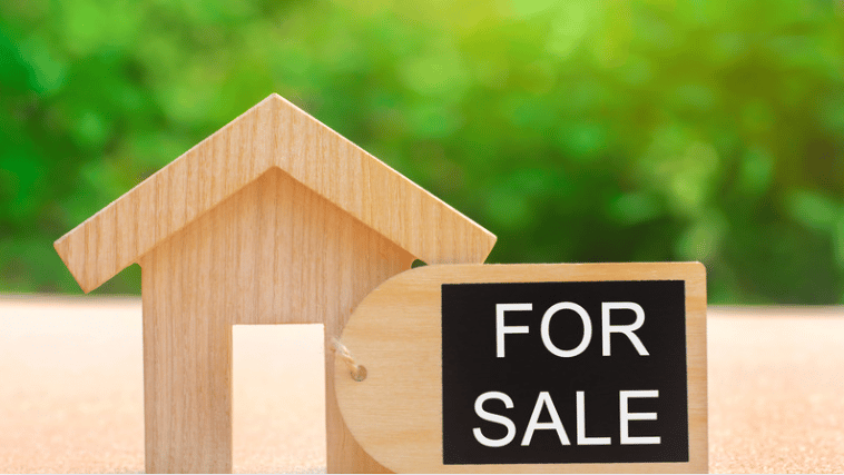 Wooden house model with a black FOR SALE sign against a blurred green background.