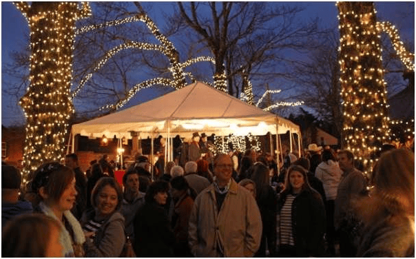 Crowded holiday gathering beneath trees wrapped in lights, with a white tent behind the guests.