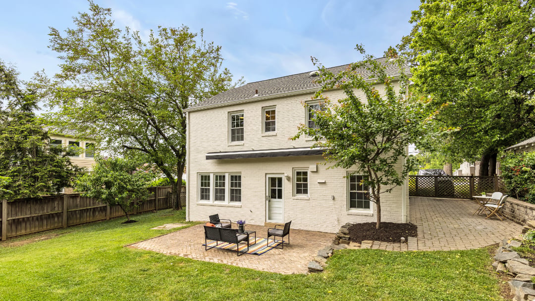 White brick home with a paved patio, outdoor seating, and a leafy fenced backyard.