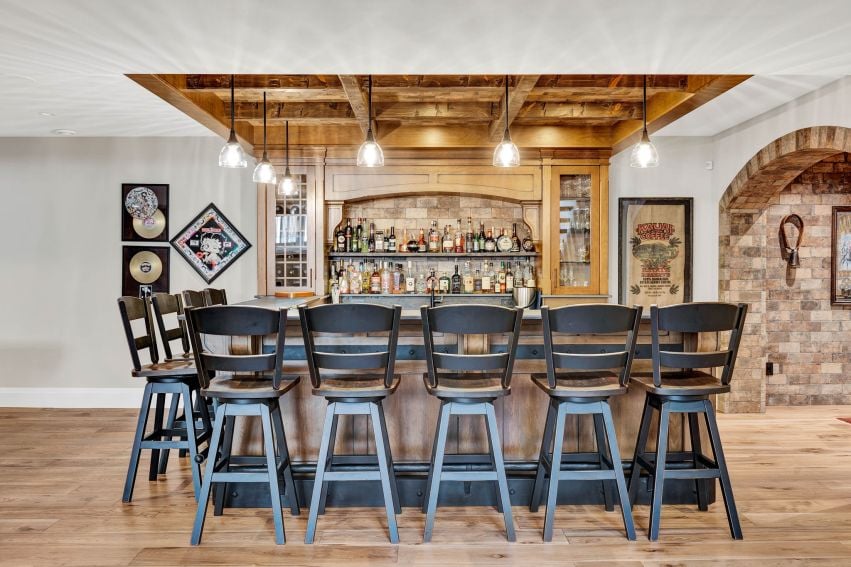 Home bar with dark stools, built-in bottle shelves, wood-beamed ceiling, and brick archway.