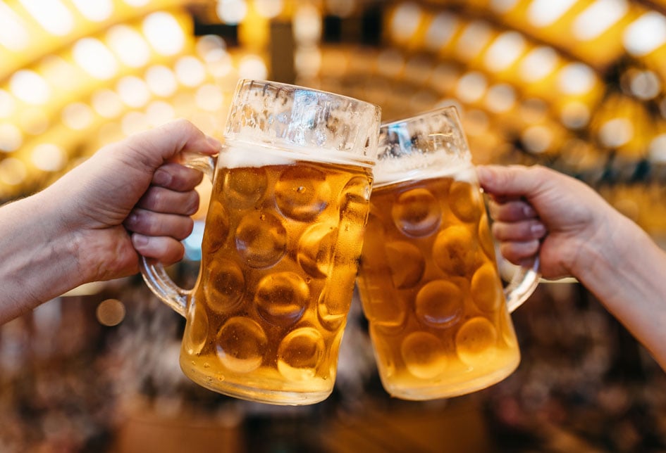 Two amber beers in glass steins clink together beneath warm Oktoberfest lights.